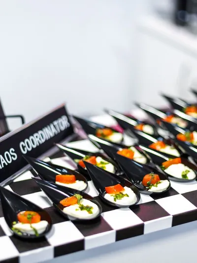 Detailný záber na elegantné predjedlá typu canapés podávané na čierno-bielom šachovnicovom podnose. Každé predjedlo je v čiernej lyžici, obsahuje bielu krémovú náplň, kúsok oranžovej zeleniny (pravdepodobne paradajky) a zelené bylinky. V pozadí je mierne rozmazaný nápis "CHAOS COORDINATOR" na čiernej tabuľke a brožúra s logom.