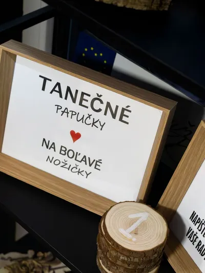 Na čiernom povrchu sú umiestnené dva drevené rámy s bielymi tabuľkami. Na ľavej tabuľke je čierny nápis „TANEČNÉ PAPUČKY NA BOĽAVÉ NOŽIČKY“ s červeným srdiečkom. Pravá tabuľka, čiastočne viditeľná, zobrazuje nápis „Rady DO MANŽELSTVA NAPÍŠTE NÁM NA PRILOŽENÉ VASE RADY A TIPY“. Medzi rámami je stoh drevených koliesok s bielou ručne maľovanou číslicou „1“. V pozadí je rozmazaná tmavomodrá dekorácia.