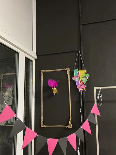 Detailný záber na tmavú stenu v salóne svadobných a spoločenských šiat. Na stene visí ozdobný zlatý rám. V ňom je zlatá nástenná lampa v tvare ruky, ktorá drží žiarovku ozdobenú sýtoružovým perím. Napravo je zavesený farebný abstraktný neónový nápis v tvare tváre. Dole je natiahnutá girlanda s ružovými a čiernymi trojuholníkovými vlajkami. Vľavo je viditeľná časť bieleho okenného rámu s odrazmi.