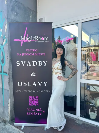 Mladá žena s tetovaniami a dlhými tmavými vlasmi pózuje v bielych, splývavých šatách so zlatými detailmi pri bannere salónu 'MagicRoom'. Banner čiernej farby s ružovým písmom propaguje 'Všetko na jednom mieste: Svadby & Oslavy' a služby ako šaty, výzdoba, darčeky. Vo výklade za ňou je vidieť svadobné šaty na figuríne.