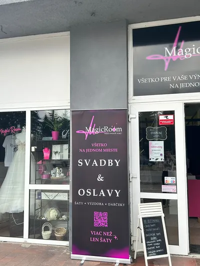 Predný pohľad na obchod MagicRoom, svadobný a spoločenský salón, s veľkým výkladom a otvorenými dverami. V ľavom výklade je vystavená biela svadobná róba a rôzne svadobné doplnky, ako šperky, košíky a dekorácie. Pred obchodom stojí ružovo-čierny roll-up banner s nápisom 'MagicRoom', 'Všetko na jednom mieste: Svadby & Oslavy, Šaty • Výzdoba • Darčeky' a QR kódom. Vedľa neho je čierna tabuľa s ponukou služieb ako organizácia svadieb a prenájom inventáru. Nad výkladom je veľký čierny nápis 'MagicRoom KED CHCEŠ VIAC' a 'VŠETKO PRE VAŠE VÝNIMOČNÉ MOMENTY NA JEDNOM MIESTE'.