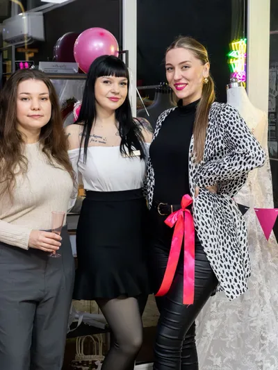Tri usmievavé ženy, pravdepodobne personál, stoja vedľa seba v modernom svadobnom a spoločenskom salóne. Žena vľavo má dlhé hnedé vlasy, béžový rebrovaný sveter, sivé nohavice a drží pohár s ružovým nápojom. Stredná žena má čierne vlasy s ofinou, biely top bez ramienok, čiernu mini-sukňu a čierne pančuchy. Žena vpravo má blond vlasy, čierny rolák, čierne kožené nohavice, čierno-bielu srsť s leopardím vzorom a výraznú fuksiovo ružovú stuhu na opasku. V pozadí sú ružové balóniky, čierno-ružová vlajková girlanda, svadobné šaty na figuríne, neónové svetlo a ružové vysoké podpätky na šachovnicovej krabici.