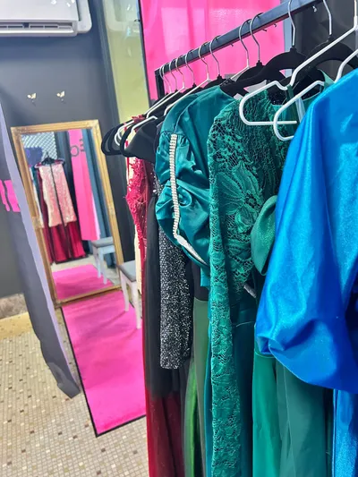 Rad elegantných spoločenských a večerných šiat v rôznych farbách a štýloch visiacich na vešiakoch. V popredí sú tyrkysové saténové šaty s naberanými rukávmi a striebornými kamienkami, smaragdovo zelené čipkované šaty a modré saténové šaty. V pozadí je zlatý rám zrkadla, ktoré odráža ružový koberec a ďalšie svetlé šaty na vešiakoch, možno svadobné, v salóne s tmavosivou stenou a kachličkovou podlahou.